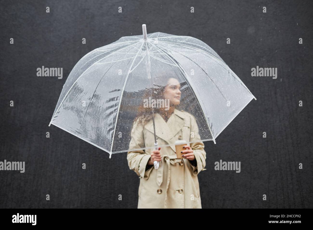 Horizontal medium shot of modern young woman wearing trench coat holding transparent umbrella and cup of coffee standing outdoors against dark gray wa Stock Photo
