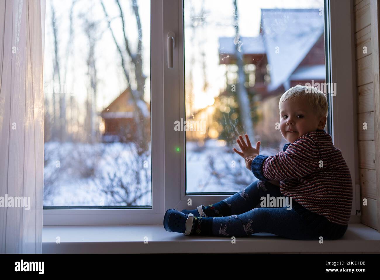 little boy stand near the window and look at the snowy street in winter, when it is very cold. Stock Photo