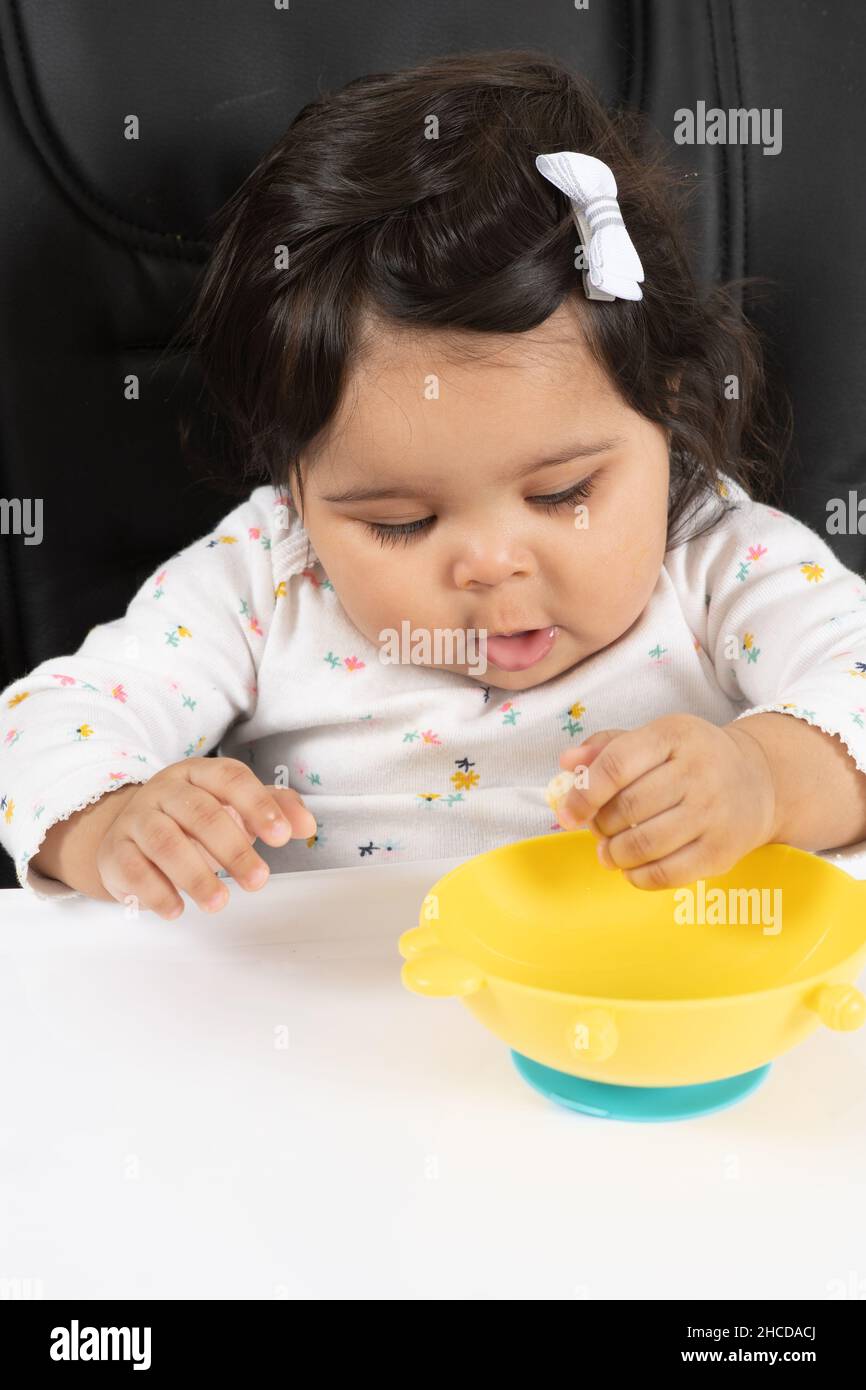 8 month old baby girl in high chair using pincer grasp to pick up piece of cereal Stock Photo