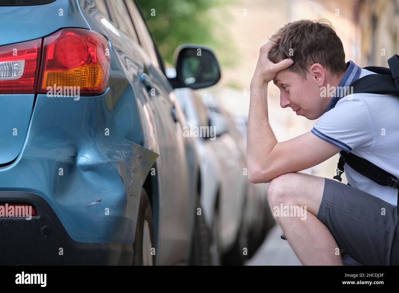 Sad young man driver sitting near his dented car looking shocked on ...