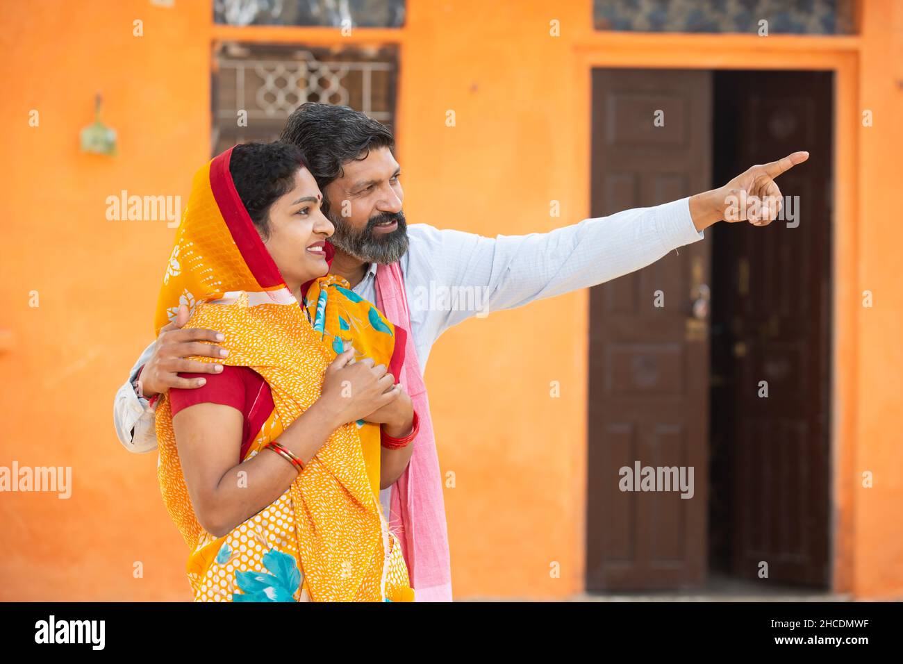 Happy beard man husband with his wife pointing at something with hand standing outside house at village, rural india, love and support. indian couple Stock Photo