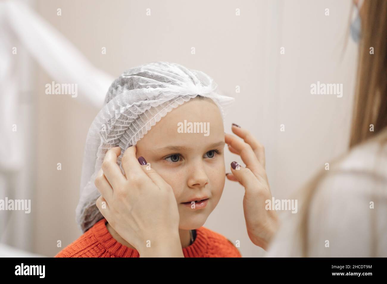 close-up of doctor hands putting white disposable medical cap on head of charming Caucasian girl in hospital ward Stock Photo