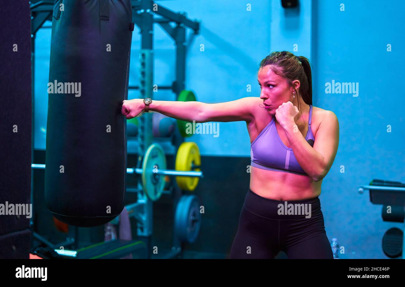 Strong ethnic female boxer punching boxing bag while training in modern