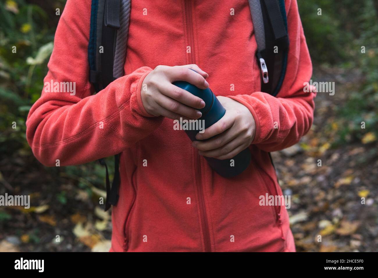 Crop anonymous female hiker with backpack opening thermos with hot drink while standing in nature with green plants during trekking Stock Photo