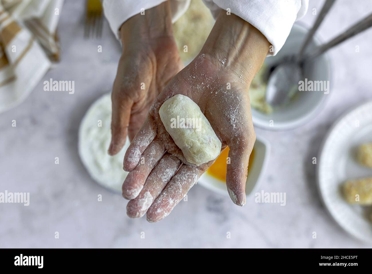 From above crop unrecognizable male cook in denim apron coating potato croquette with flour while standing at messy table in light kitchen Stock Photo