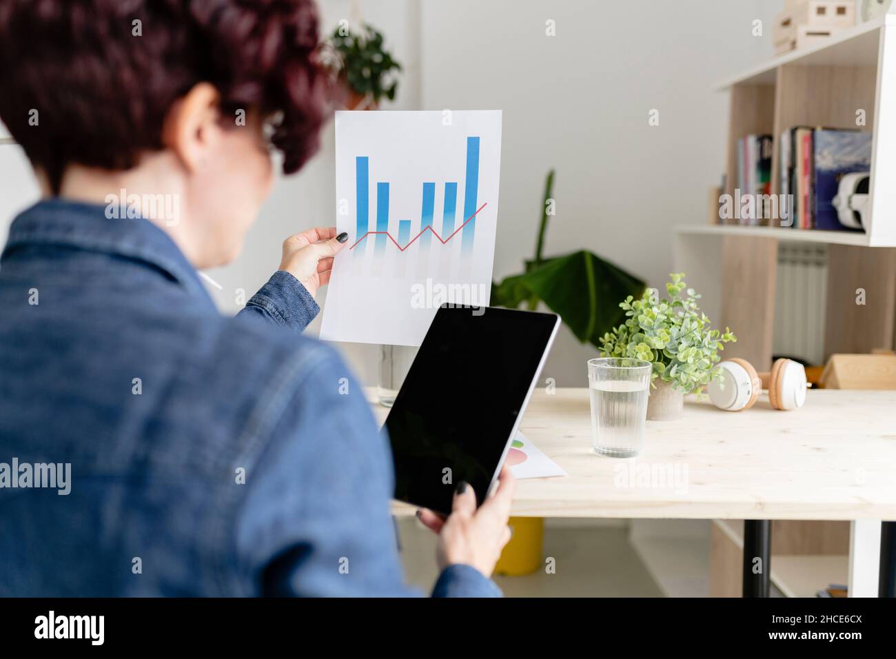 Back view of crop anonymous female with modern tablet with black screen sitting at table while analyzing charts on paper in home office Stock Photo