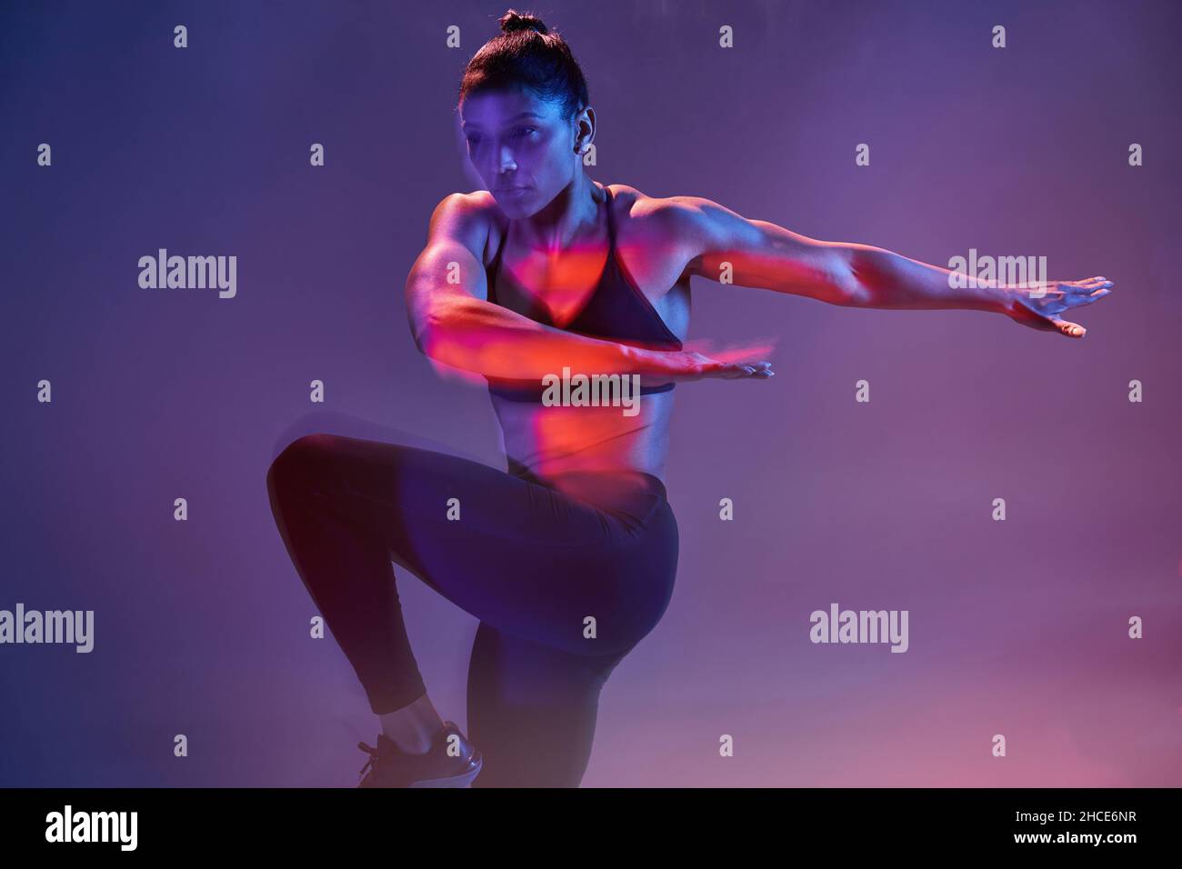 Fit young female in activewear performing exercise while looking away in dark studio with purple neon illumination Stock Photo