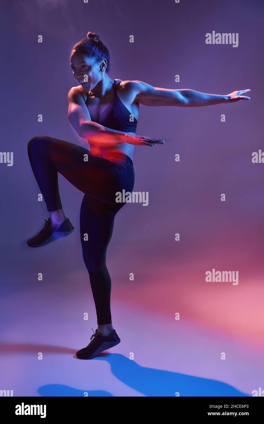 Fit young female in activewear performing exercise while looking away in dark studio with purple neon illumination Stock Photo