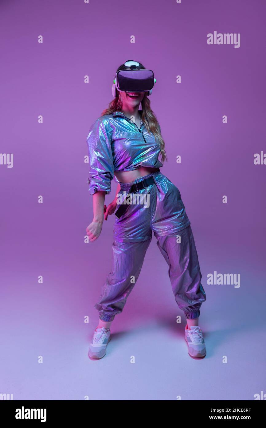 Full body of cheerful female with long hair in trendy outfit and VR glasses dancing in studio with purple neon illumination Stock Photo