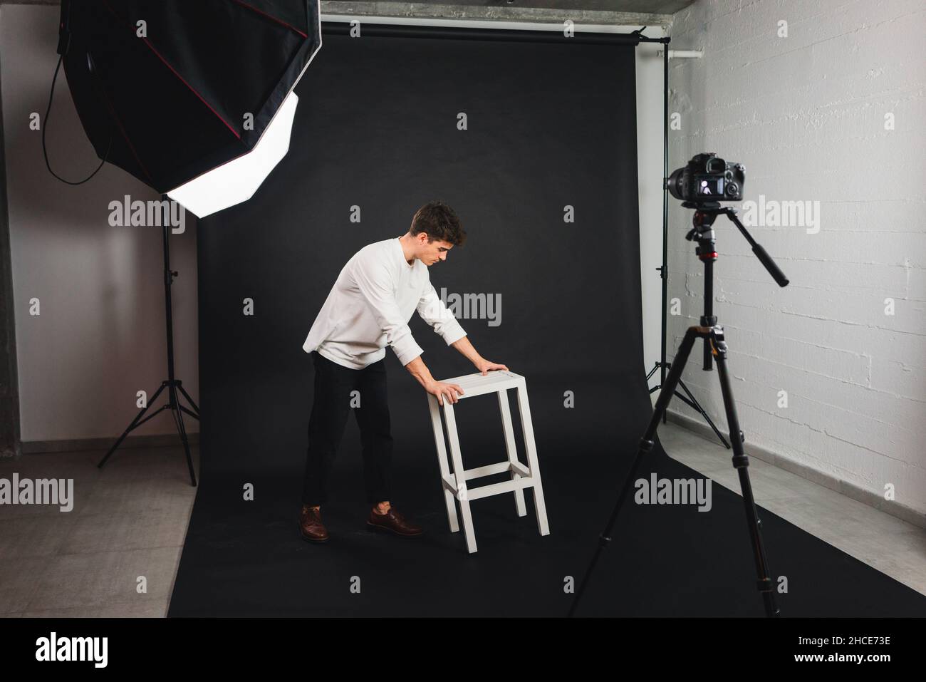 Full body of guy in casual clothes carrying white wooden chair on black background in professional photo studio Stock Photo