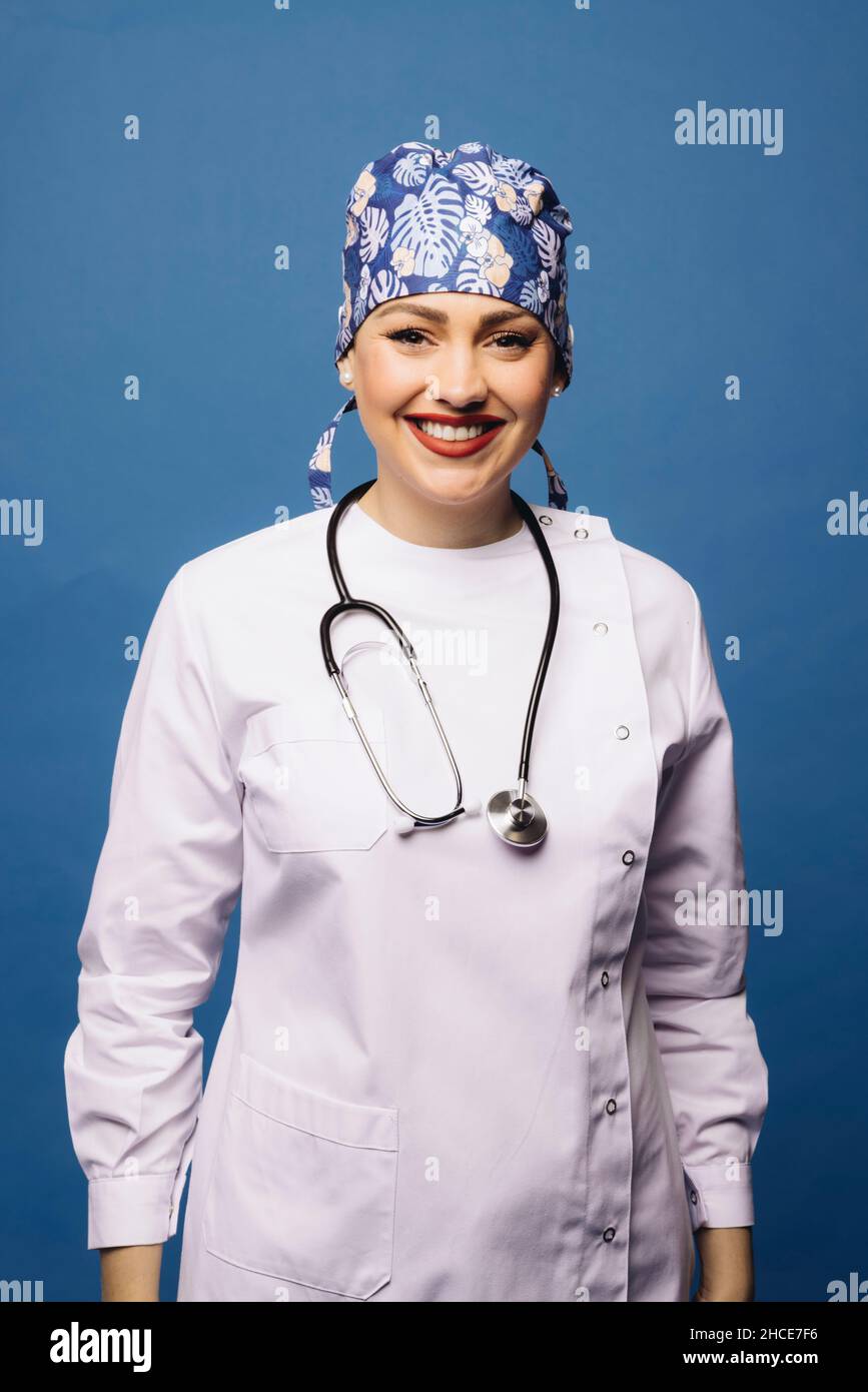 Positive doctor Woman in white uniform robe and bandana with stethoscope looking at camera on blue background Stock Photo