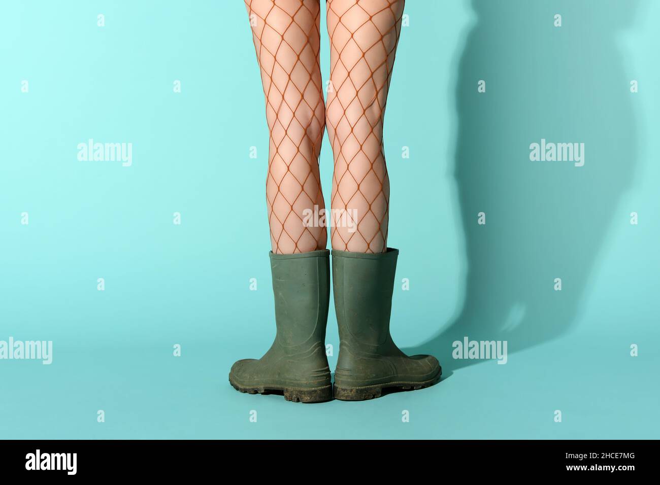 Crop unrecognizable female model wearing fishnet stockings and dirty rubber boots standing in blue studio Stock Photo