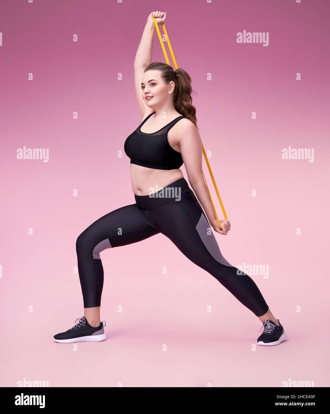 Strong girl working with resistance band. Photo of model with curvy figure in fashionable sportswear on pink background. Sports motivation and healthy Stock Photo
