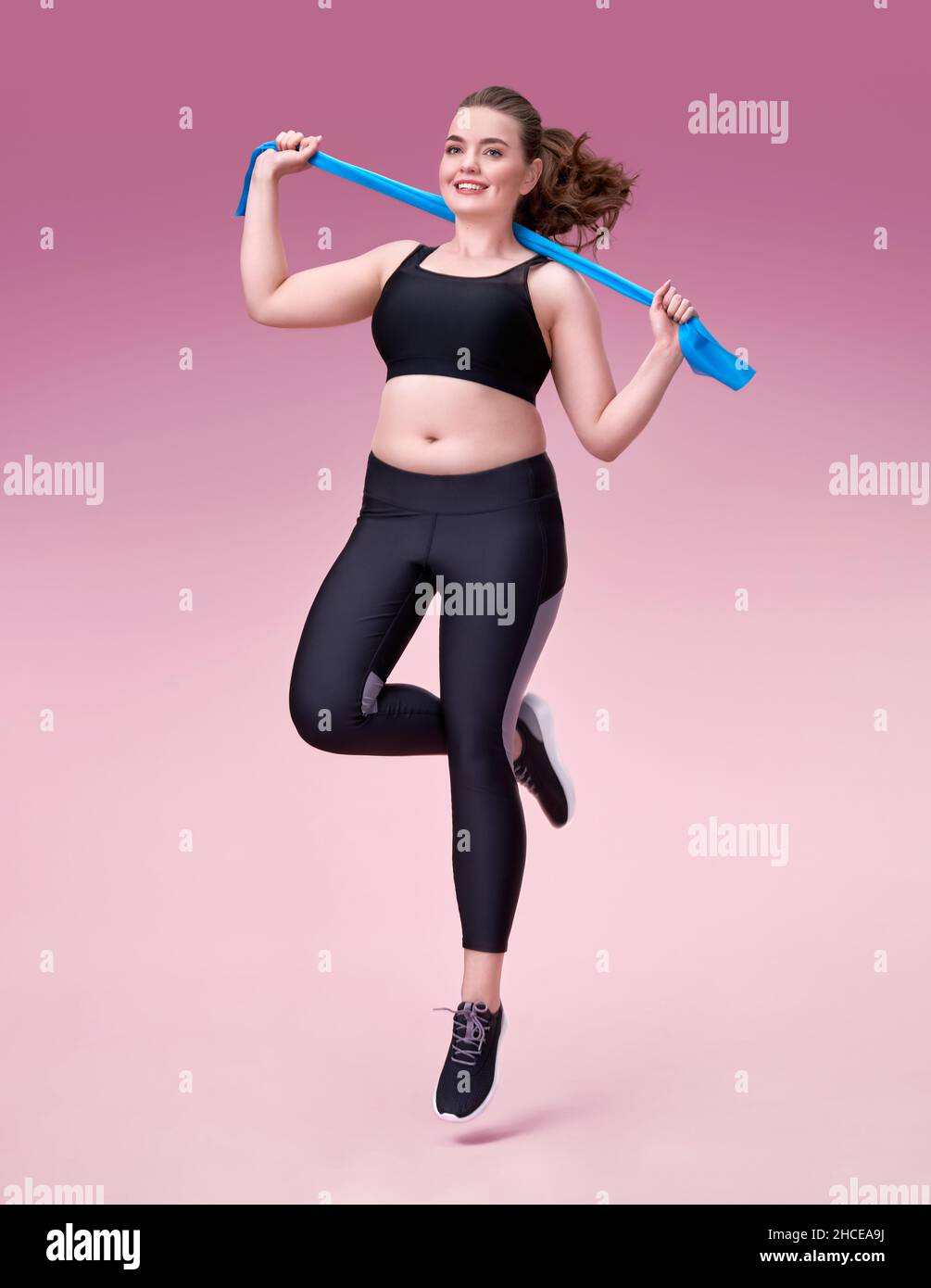 Sporty girl with resistance band in motion. Photo of model with curvy figure in fashionable sportswear on pink background. Dynamic movement. Sports mo Stock Photo