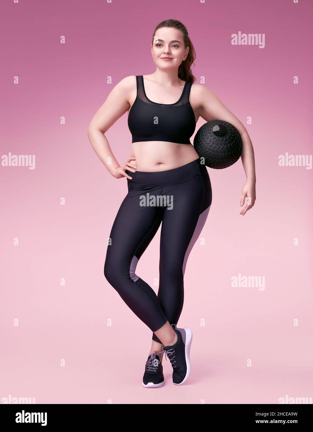 Sporty girl resting after workout with medicine ball. Photo of model with curvy figure in fashionable sportswear on pink background. Sports motivation Stock Photo