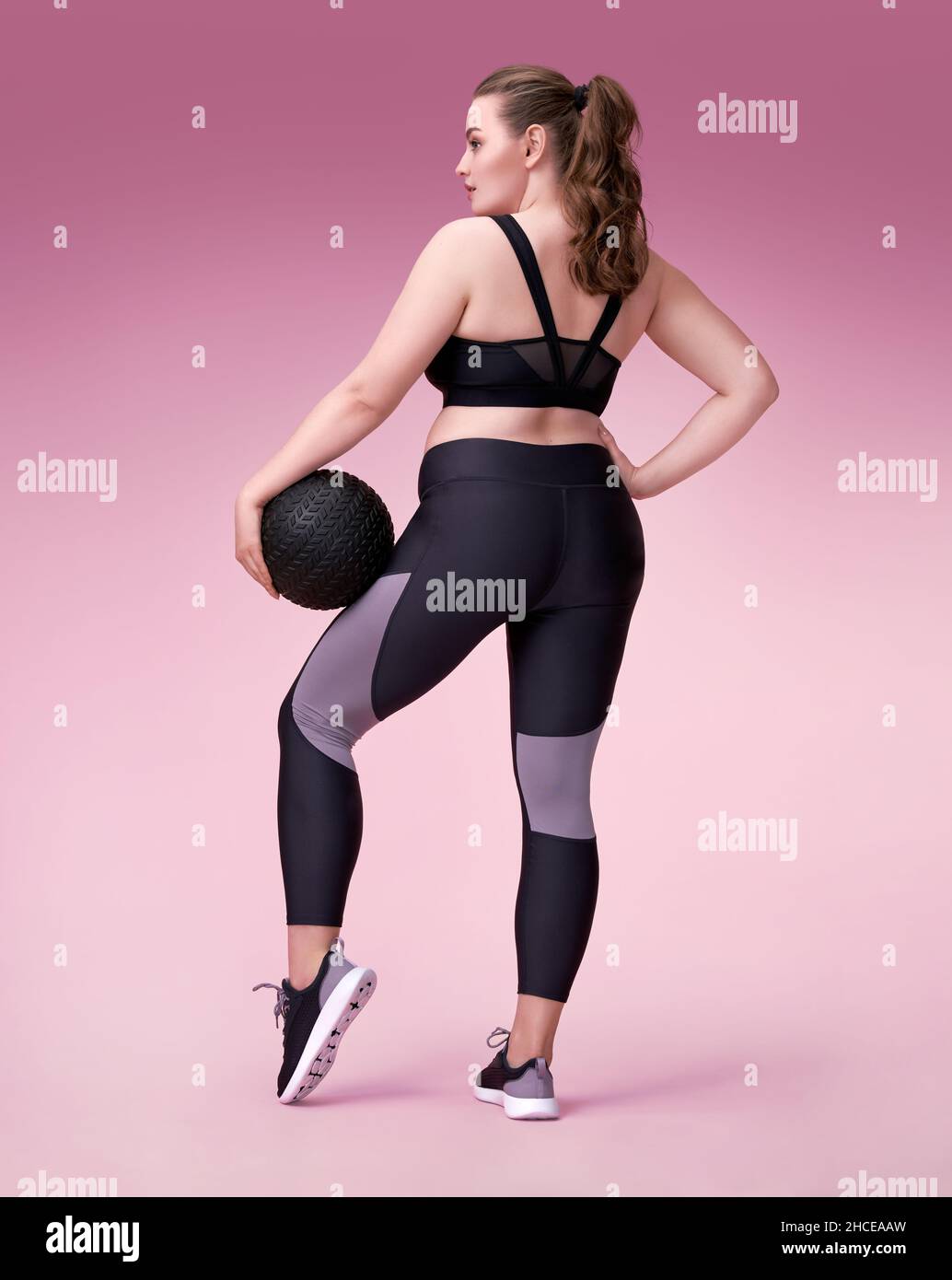 Sporty woman with medicine ball. Photo of model with curvy figure in fashionable sportswear on pink background. Sports motivation and healthy lifestyl Stock Photo