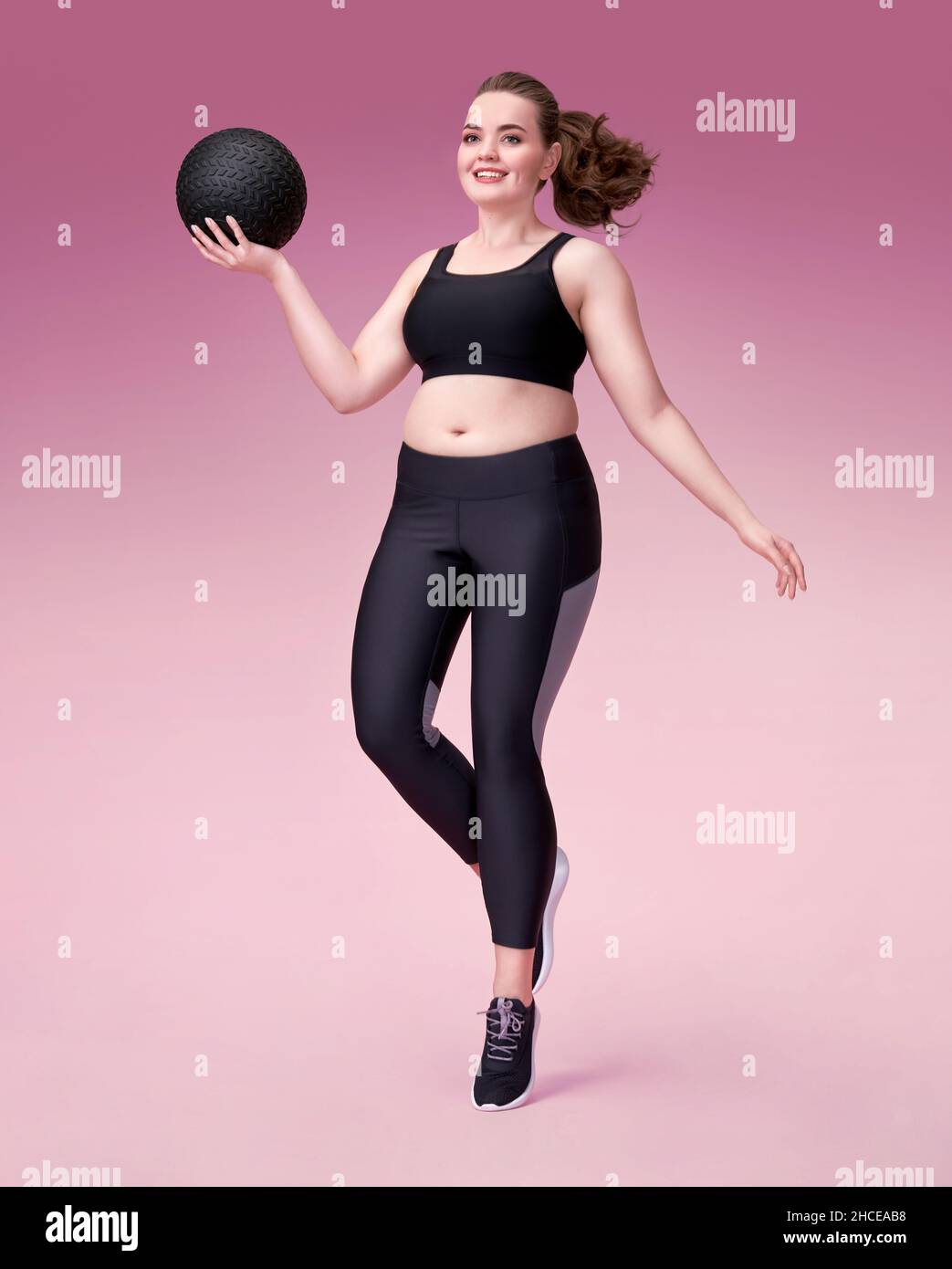 Sporty girl with medicine ball in motion. Photo of model with curvy figure in fashionable sportswear on pink background. Dynamic movement. Sports moti Stock Photo
