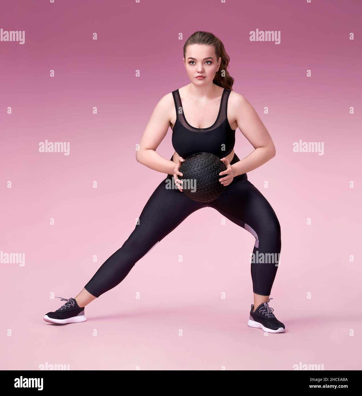 Sporty woman doing exercise with medicine ball. Photo of model with curvy figure in fashionable sportswear on pink background. Sports motivation and h Stock Photo