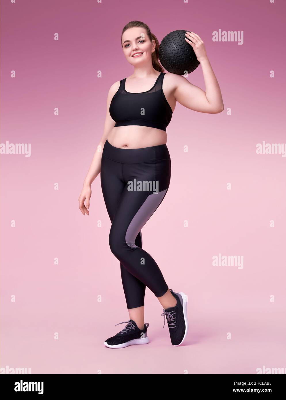 Sporty woman with medicine ball on her shoulder. Photo of model with curvy figure in fashionable sportswear on pink background. Sports motivation and Stock Photo