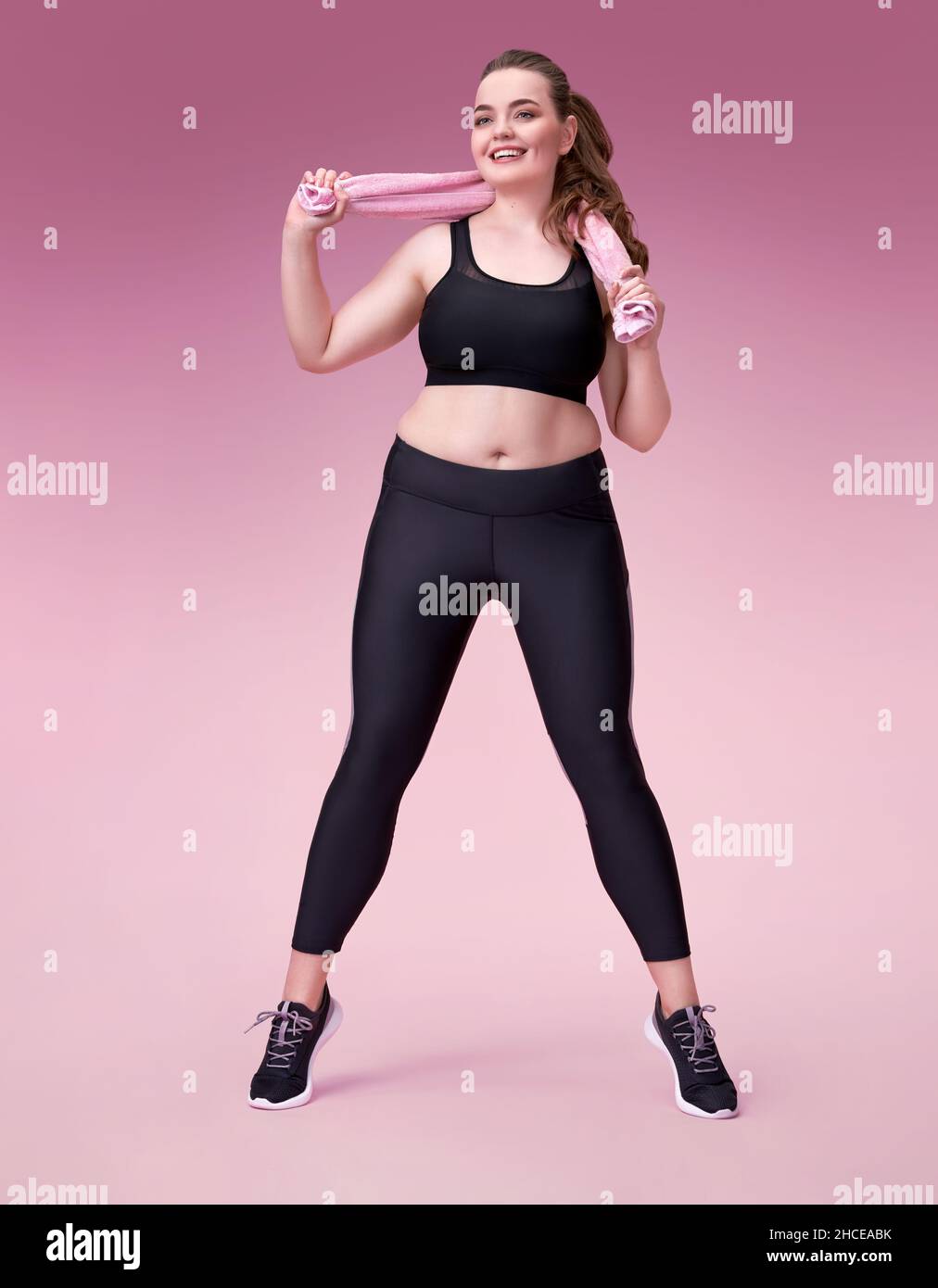 Sporty girl with towel after training. Photo of model with curvy figure in fashionable sportswear on pink background. Sports motivation and healthy li Stock Photo
