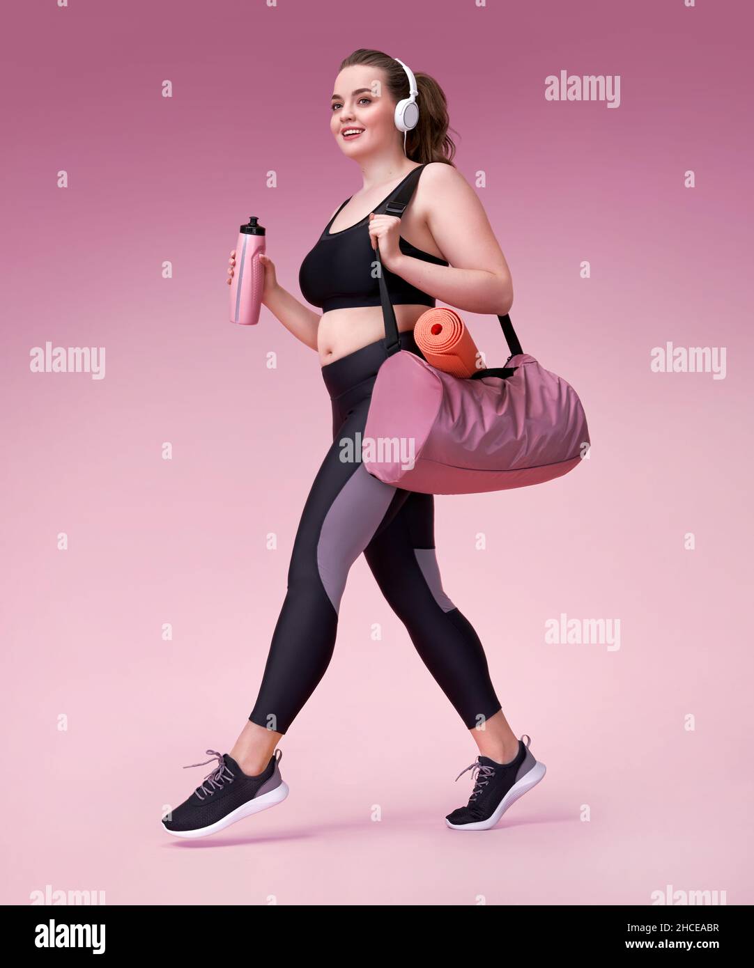Go to training! Smiling sporty woman with sports bag and shaker. Photo of model with curvy figure in fashionable sportswear on pink background. Sports Stock Photo