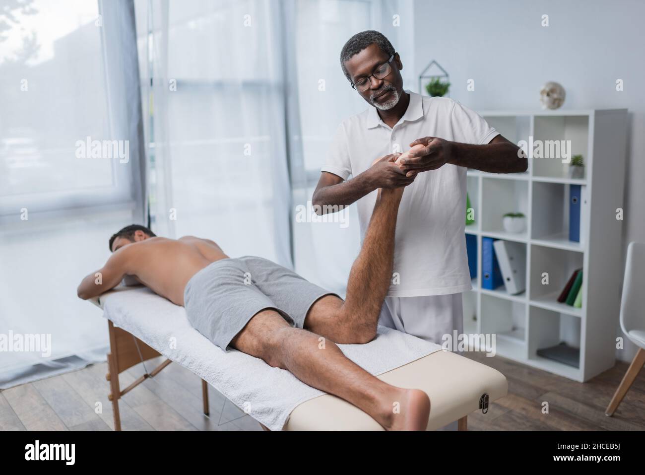 african american rehabilitologist massaging foot of young man in clinic Stock Photo