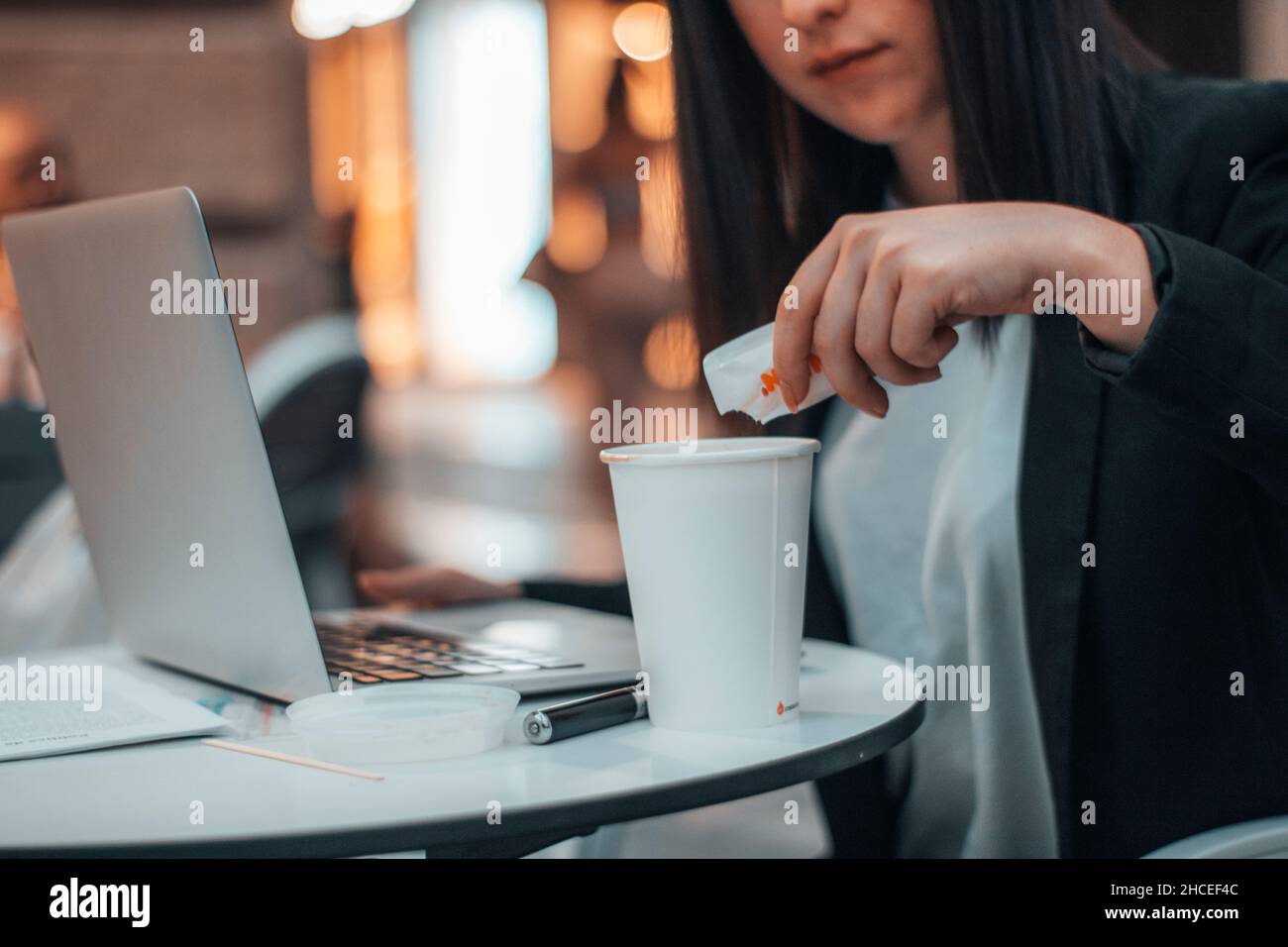 Young manager worker pouring sugar into coffee in a cafeteria in the city square while working with her laptop Stock Photo