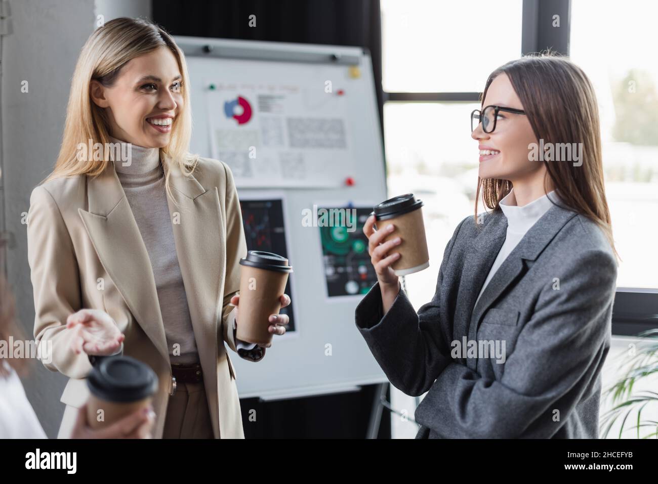 cheerful businesswomen with paper cups talking near blurred flip chart ...