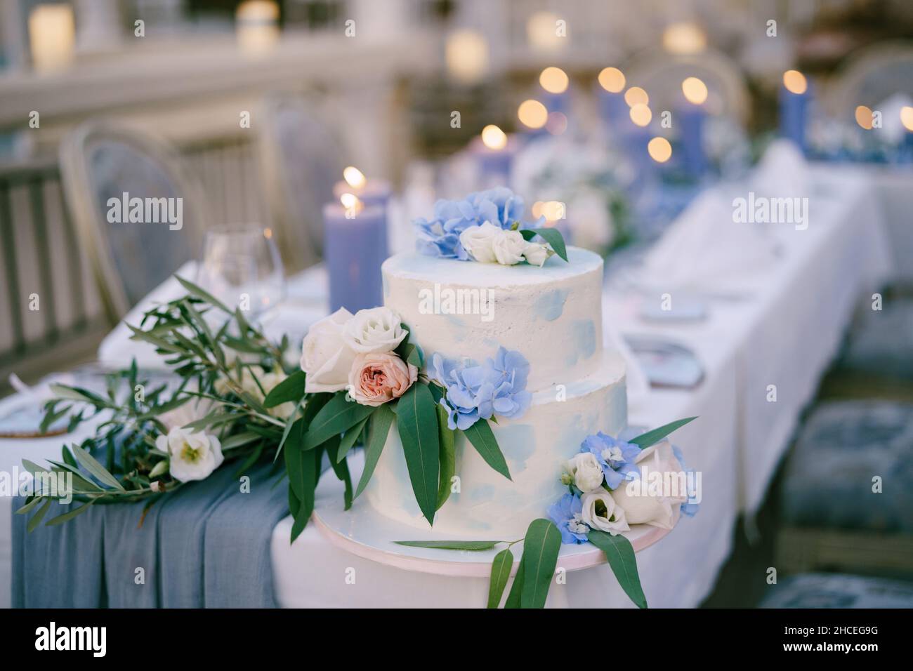 White bunk wedding cake decorated with flowers and leaves stands on a table with candles Stock Photo