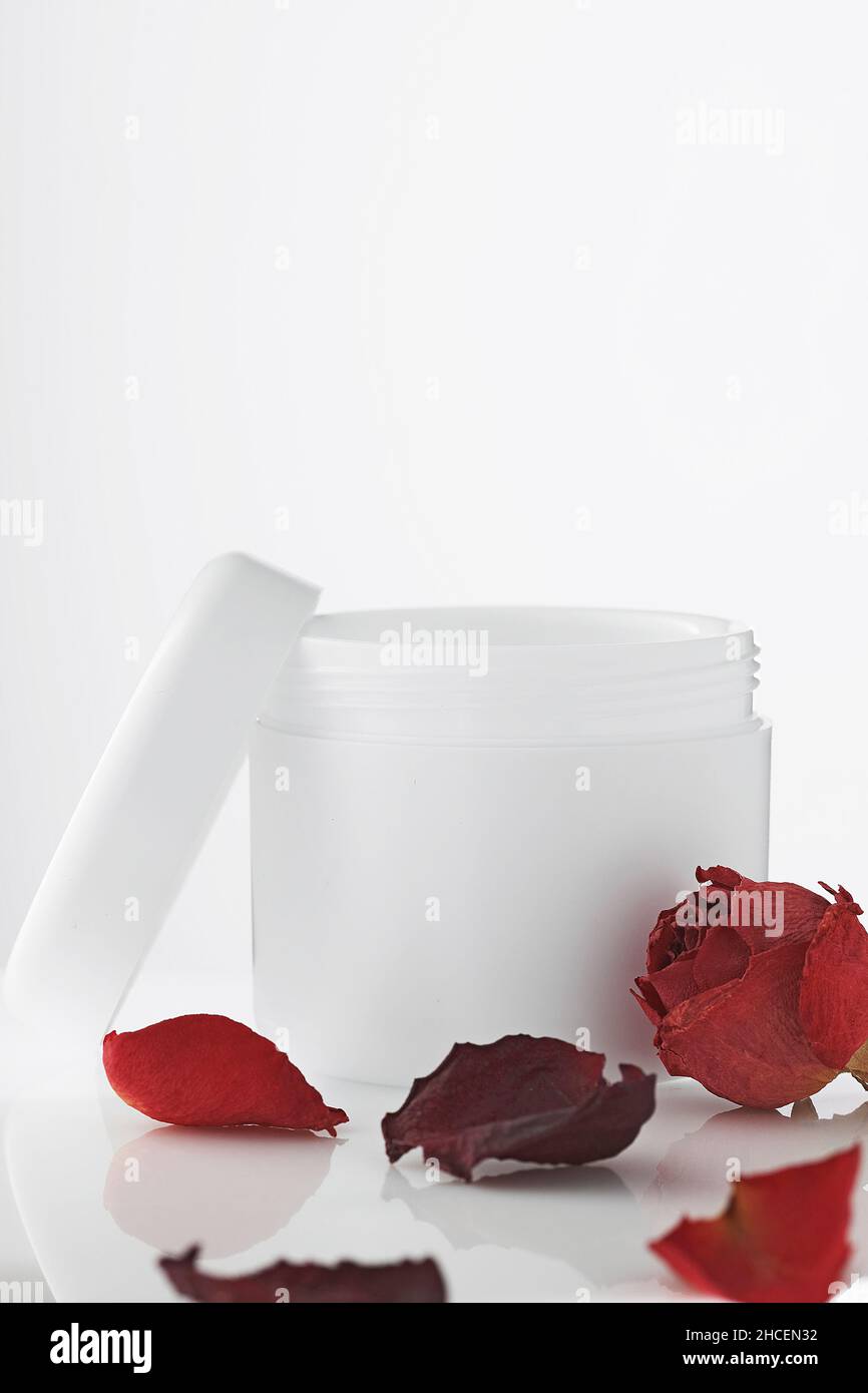 Empty white plastic jar with lid open on light background. Petals of red roses. Rejuvenating cosmetics. Care product for skin of body and face. Layout for logo. Cosmetology and spa. Stock Photo