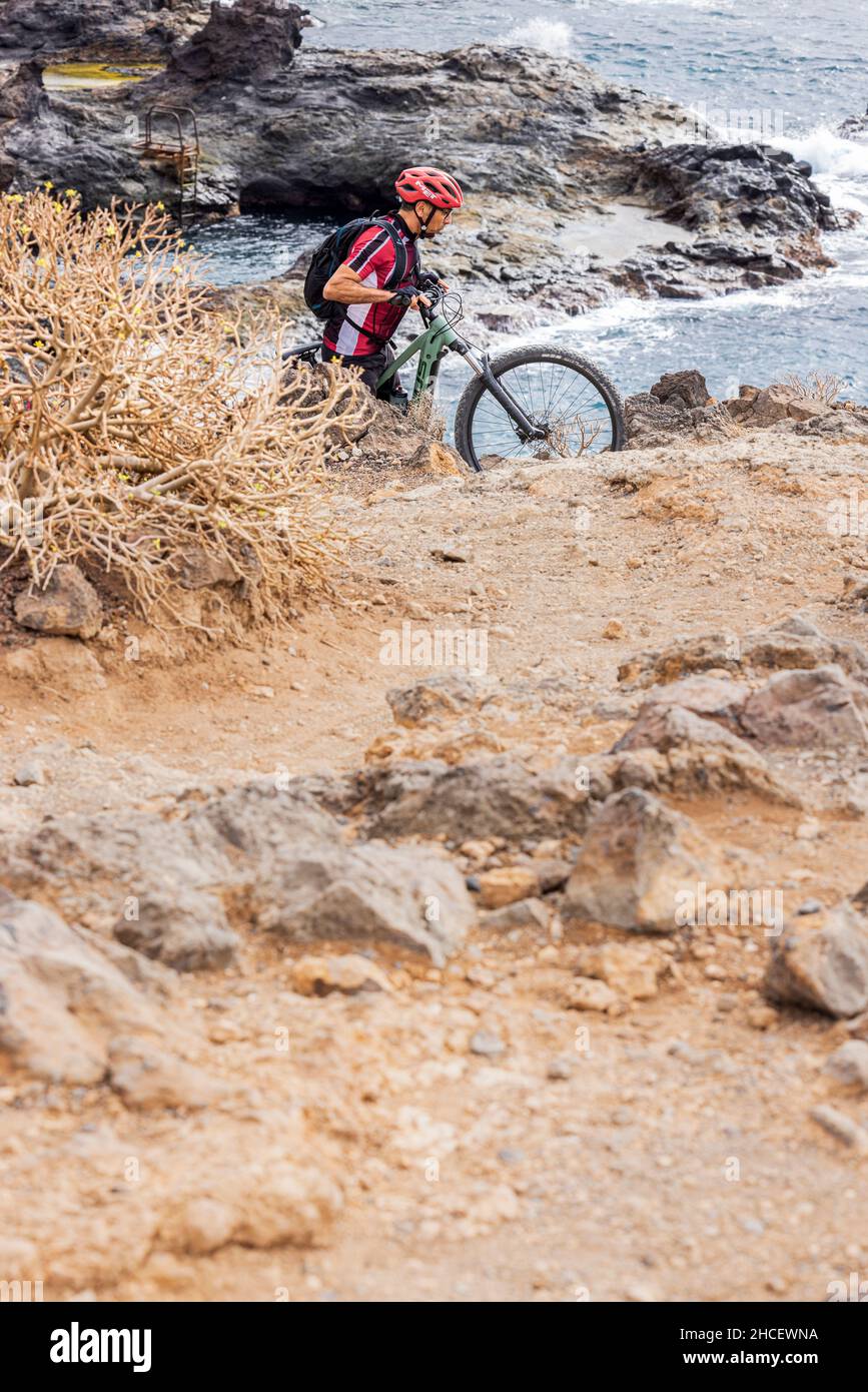 Mountain biker pushes his e bike up a steep slope on rough ground by the coast near Los Abrigos, Tenerife, Canary Islands, Spain Stock Photo