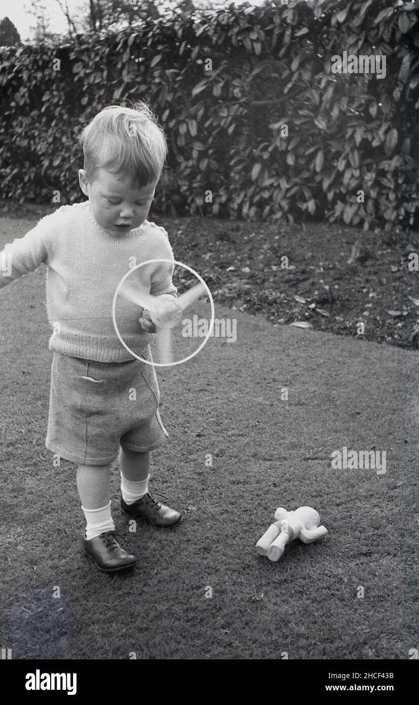 1950s, hisotrical, outside in a garden, a little boy in a woollen top and shorts, playing with his spinning top toy, England, UK. Spining toys were a popular play item in this decade and came in a variety of forms and materials.  The toy here is a light handheld model, where a string was pulled from the handle to spin the plastic circular top around. Stock Photo