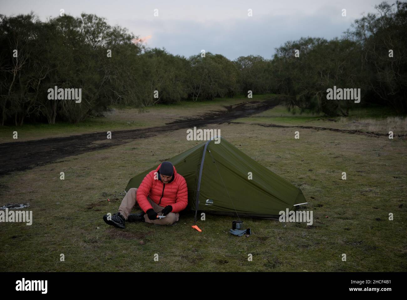 wild camp and survival skills in outdoor activity a man is sharpening a wooden branch sitting in front of tent Stock Photo
