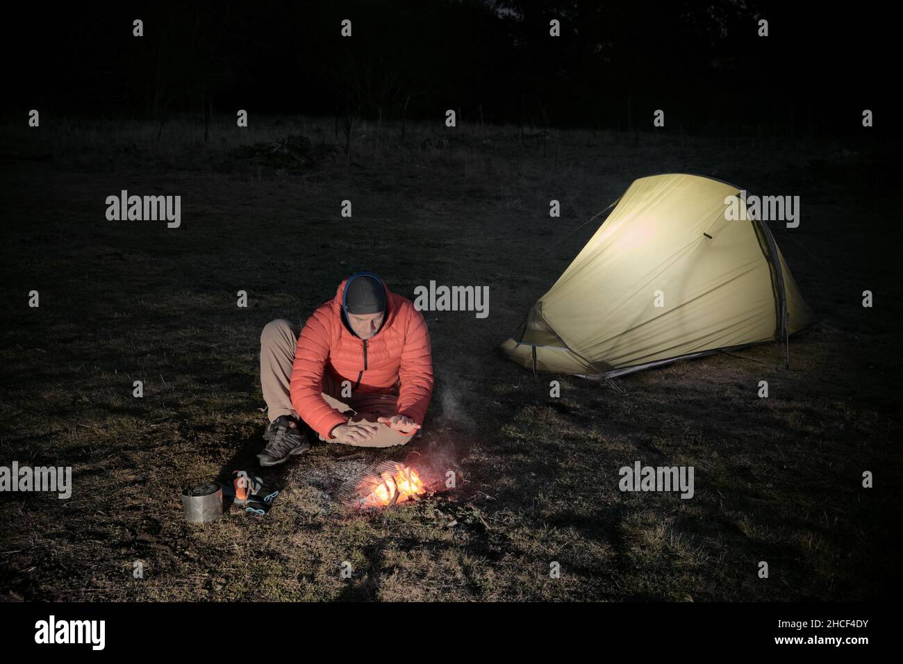 wild camping and survival skills in outdoor activity a man heats hands on camp fire sitting nearby the illuminated tent Stock Photo