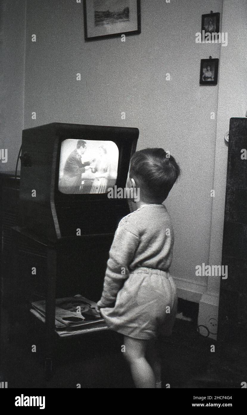 Late 1950s, historical, a little boy standing up close to a small television set of the era watching a programme, England, UK. In 1950, a television set was a luxury item, with only around 350,000 British households owning one. but by the end of the decade, nearly 75% of the population had television.K. Copies of the popular female magazine, 'Woman' are lying underneath the TV. Stock Photo
