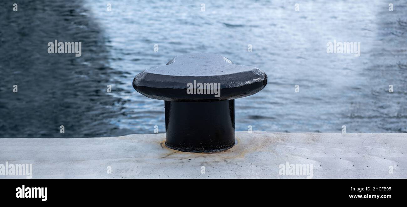 Black bollard on harbor pier, rippled sea water background, copy space. Heavy metal post anchored on quay concrete floor Stock Photo