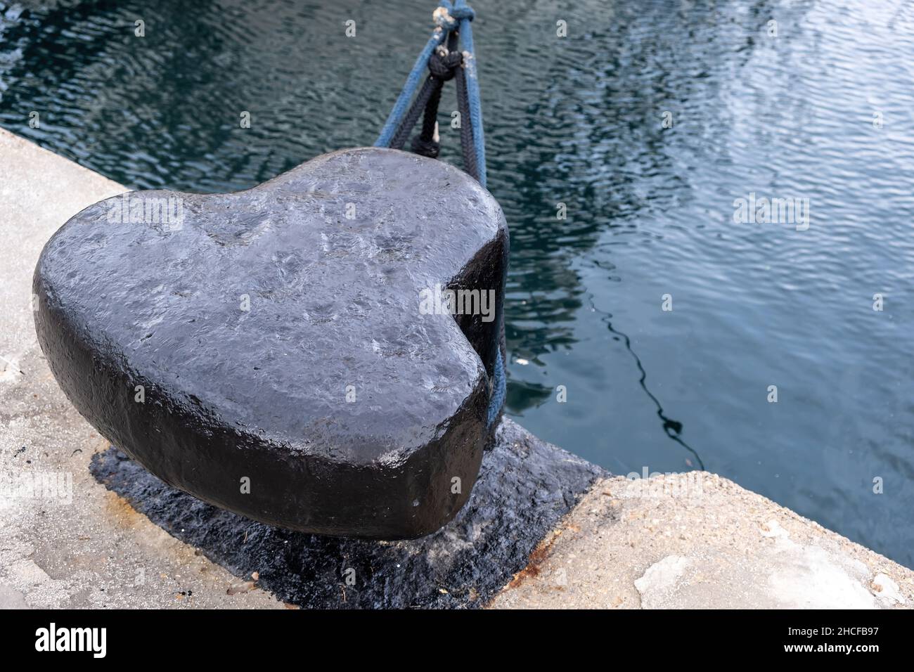 Ship mooring rope around a port bollard on harbor pier, rippled sea water background, copy space. Black metal post anchored on quay concrete floor Stock Photo