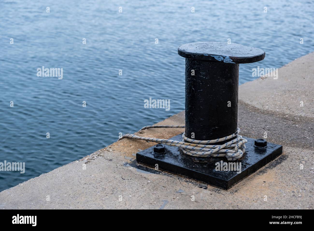 Ship mooring rope around a port bollard on harbor pier, rippled sea water background, copy space. Black metal post anchored on quay concrete floor Stock Photo