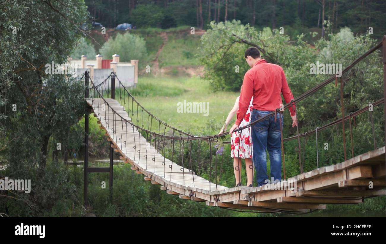 Young lovers on a rope bridge across the river. A couple of hikers traversing a swing bridge. Clip. young couple in love walking on suspended wooden b Stock Photo