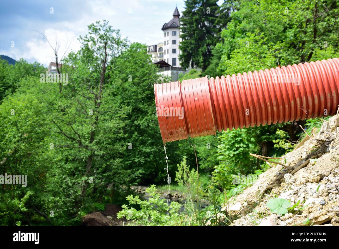 A thin stream of water flows from a red plastic sewer pipe against a blurred background of trees and a building Stock Photo