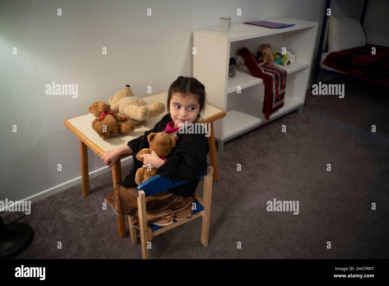 Austin Texas USA, December 2021: An Afghan child cuddles a stuffed bear and sits in a child-sized chair in the bedroom of her family's new apartment near downtown. Refugees fleeing turmoil in Afghanistan continue to be resettled in Texas with many single men and large families with children moving into apartments across Austin. Faith-based and other non-profit groups have stepped up to help coordinate efforts in furnishing homes for hundreds of refugees. ©Bob Daemmrich Stock Photo