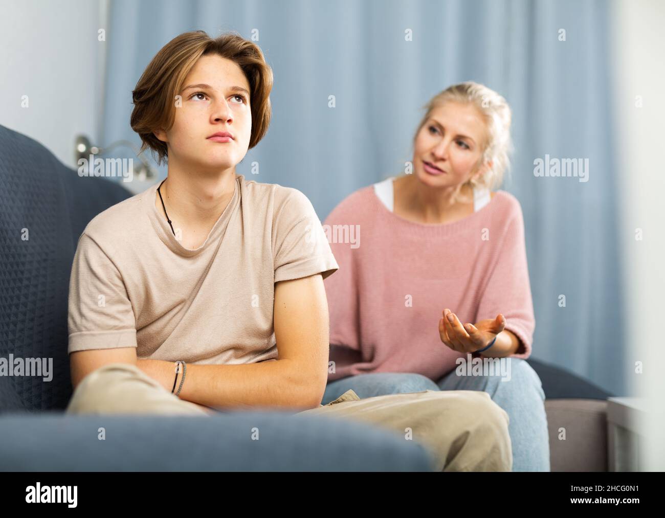 Upset and offended teenager sitting on sofa while mother soothing him Stock Photo