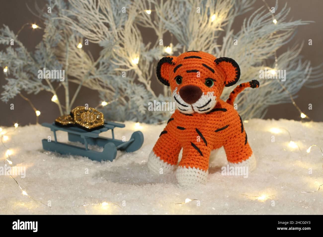 A knitted woolen tiger cub sits next to toy light blue trees with lights and toy sleds with coins on artificial white snow on a gray background. Close Stock Photo