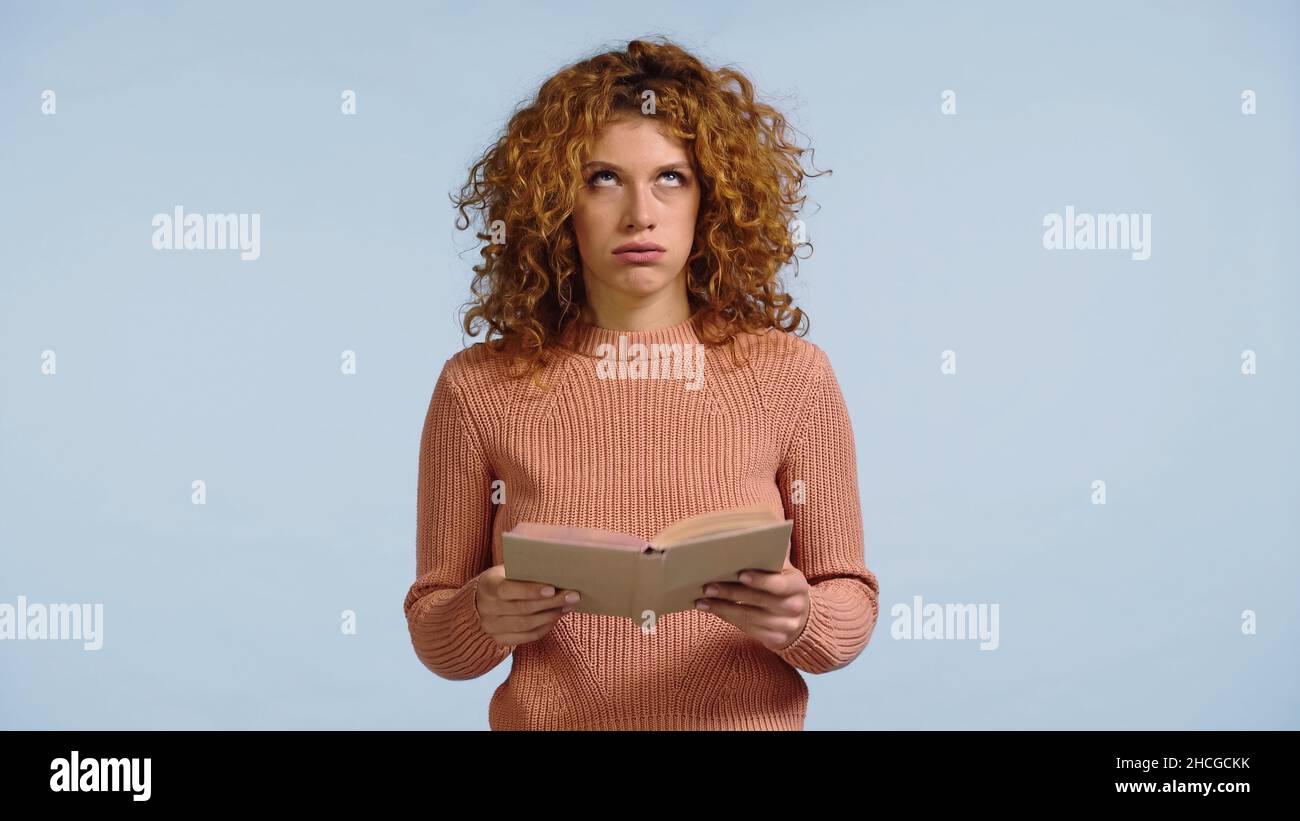 exhausted redhead woman with book looking up and puffing cheeks isolated on blue Stock Photo