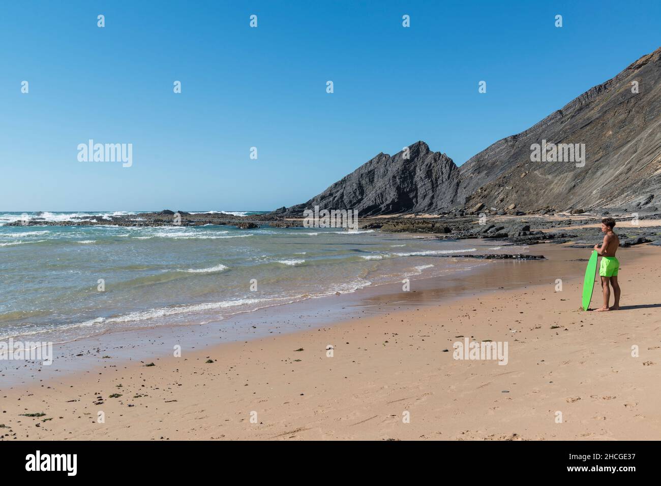 Young man in fluorescent green swimming trunks holding a surfboard of the same color looks at the sea at Praia da Amoreira beach, Algarve, Portugal. Stock Photo