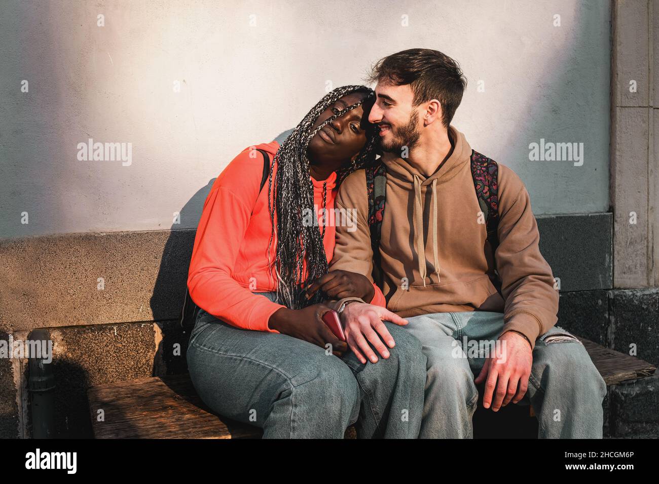 A couple of young people in love of different nationalities and cultures are resting in the street sitting on a bench Stock Photo