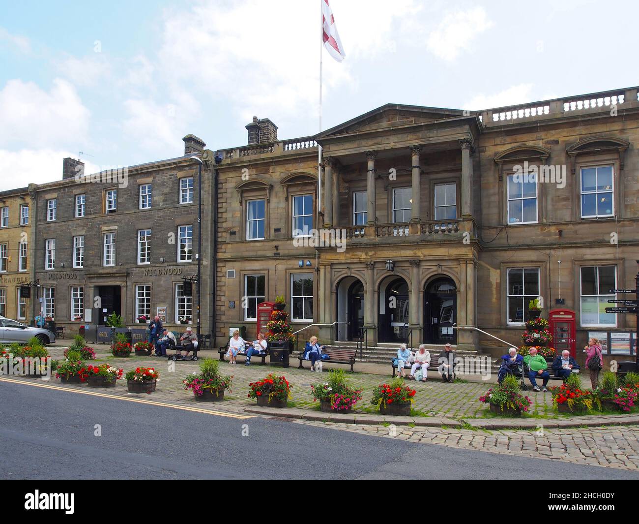 Senior citizens enjoying the sunshine sitting on benches outside Skipton town hall, North Yorkshire, England, with flowers in tubs in the foreground. Stock Photo