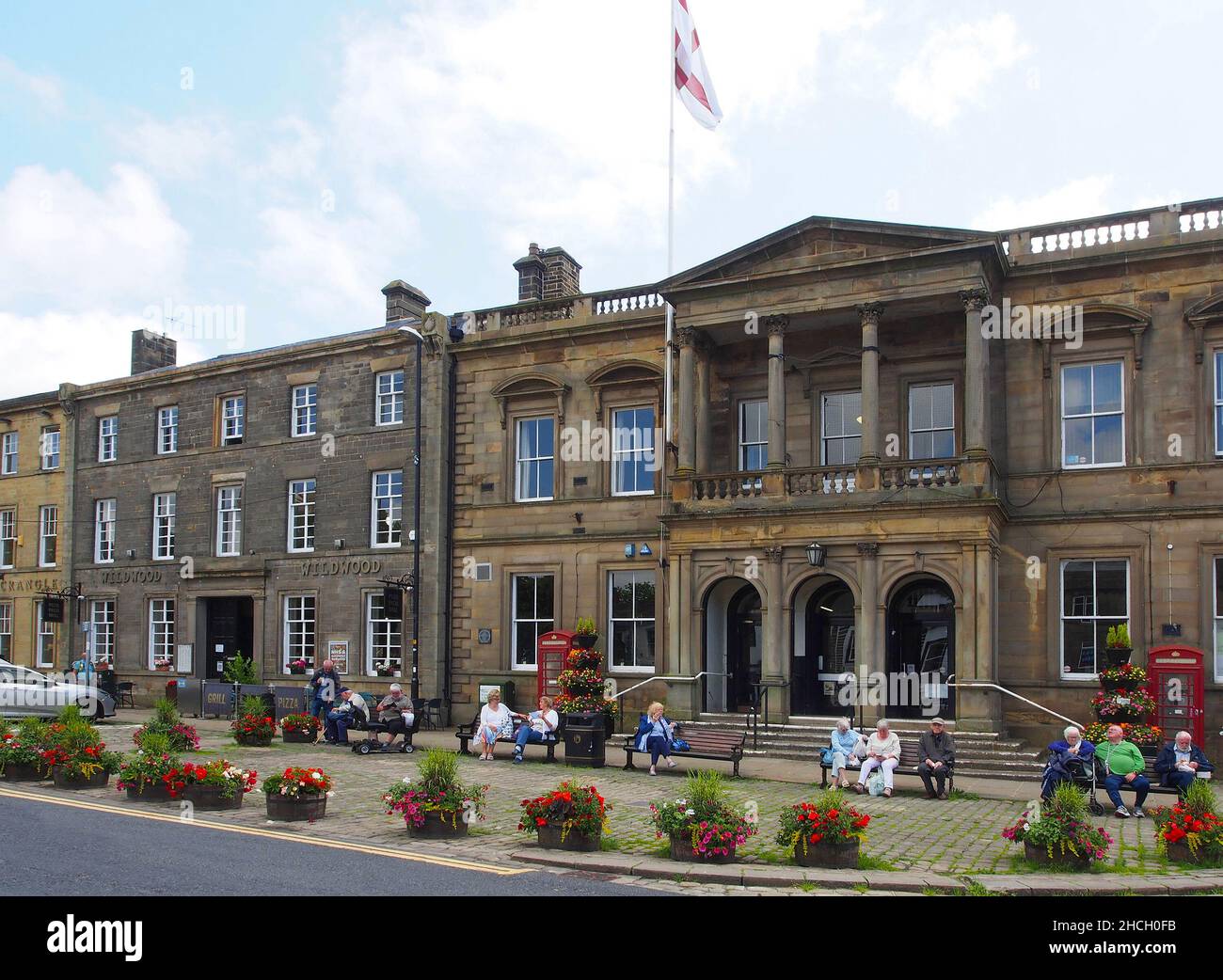Senior citizens enjoying the sunshine sitting on benches outside Skipton town hall, North Yorkshire, England, with flowers in tubs in the foreground. Stock Photo