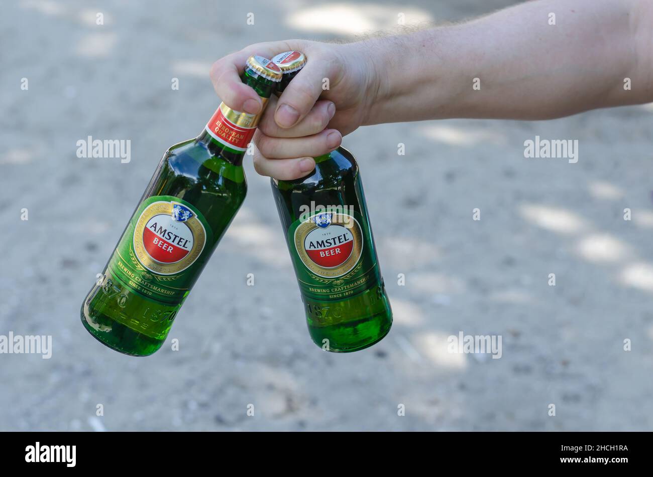 A hand holds two bottles of Amstel beer against the sand. Adult male with green bottles of alcoholic beverage. Nikolaev, Ukraine - 07 13 2021 Stock Photo