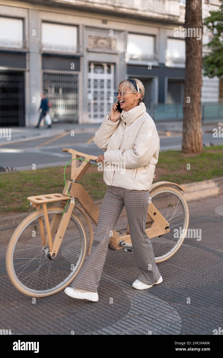 Optimistic woman in outerwear laughing at joke while walking near wooden eco bike and talking on cellphone on city street. Stock Photo
